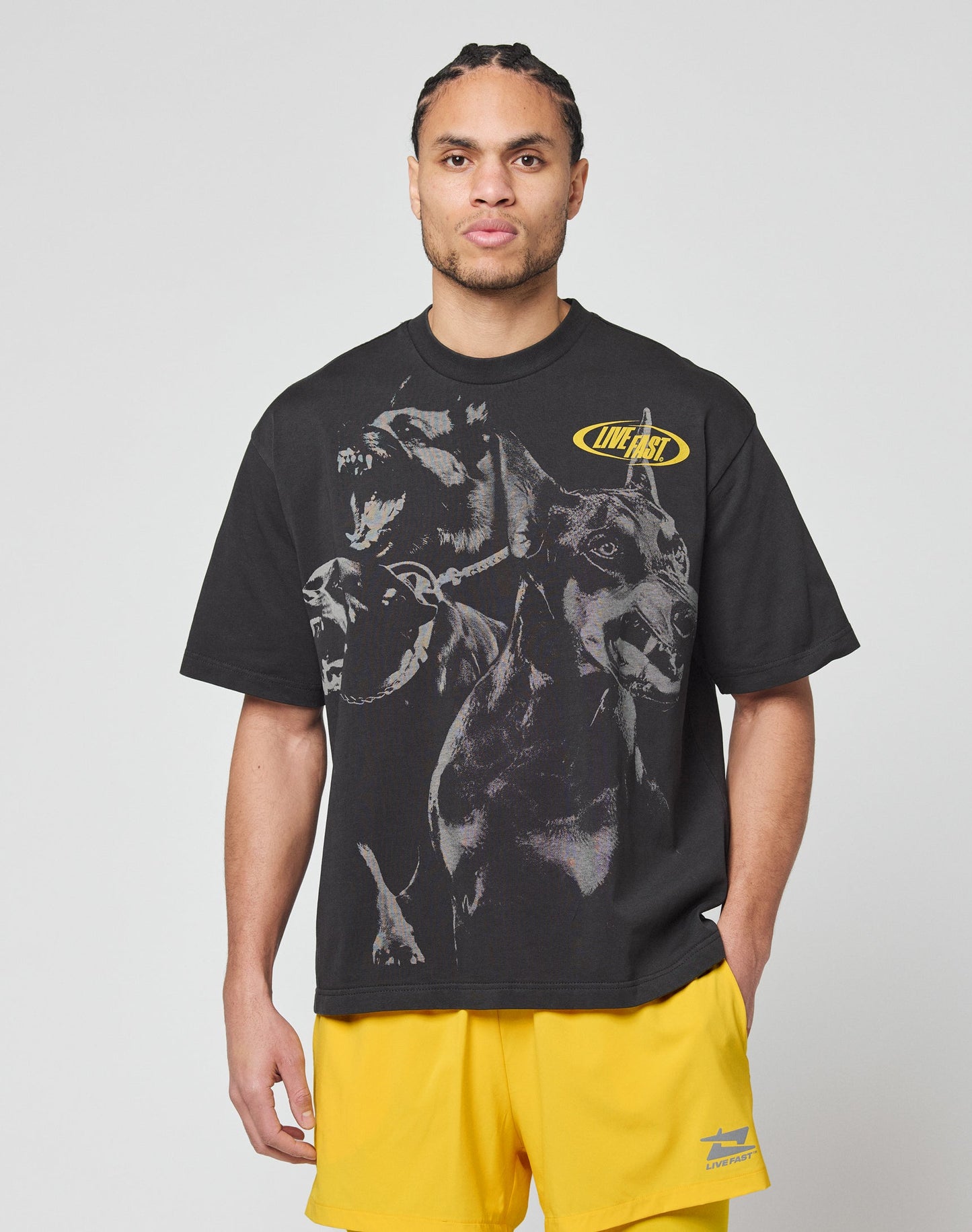A man with braided hair wears the LIVE FAST DIE YOUNG LFDY Bark Tee, a black, vintage wash shirt with large dog graphics and yellow LIVE FAST text. He pairs it with bright yellow athletic shorts against a plain gray background.
