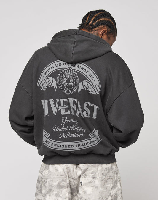 A person with braided hair faces away, wearing the oversize black LFDY Loyal Hooded Zip by LIVE FAST DIE YOUNG in 100% cotton with vintage wash, bold white graphics, and banners reading “Live Fast,” countries, and “With us or against us.”.