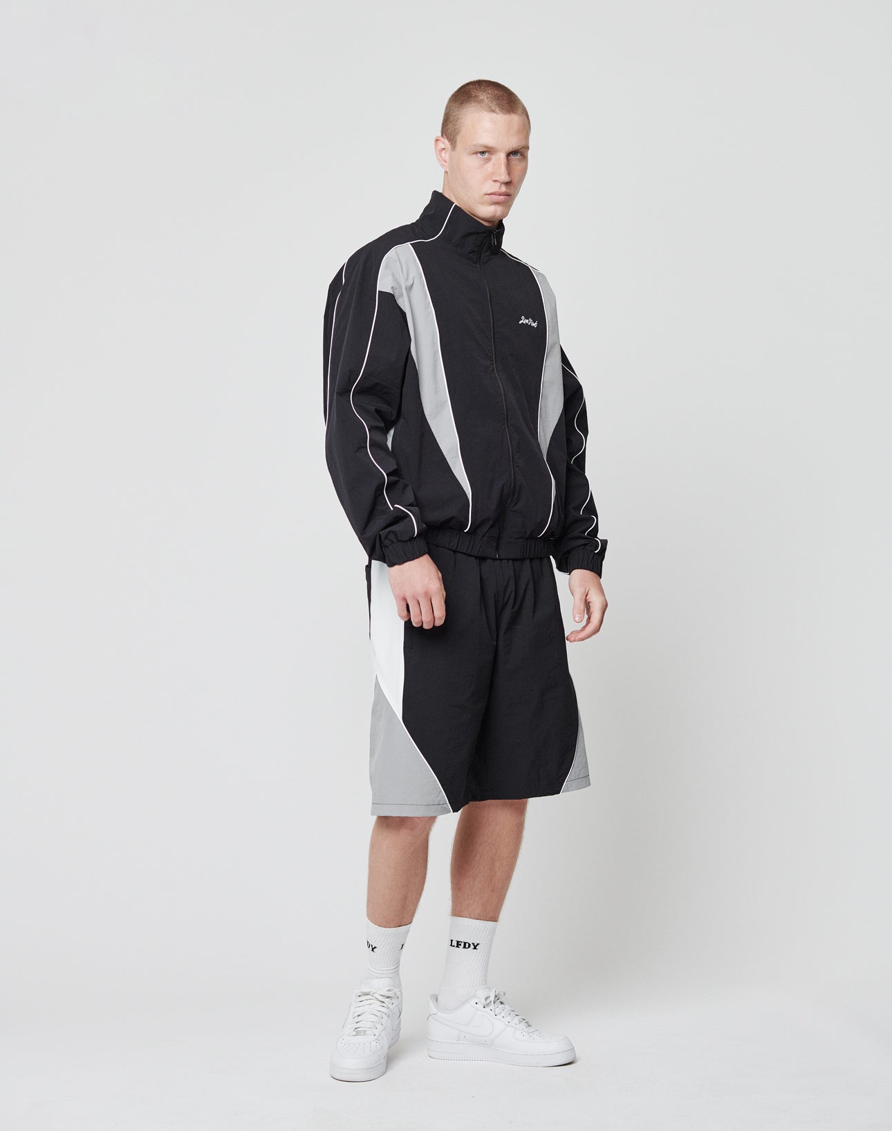 A man wearing LIVE FAST DIE YOUNG Basic Nylon Trackshorts, a matching black and gray nylon jacket with elastisches Bündchen, white socks, and sneakers stands against a plain white background, looking directly at the camera with a neutral expression.
