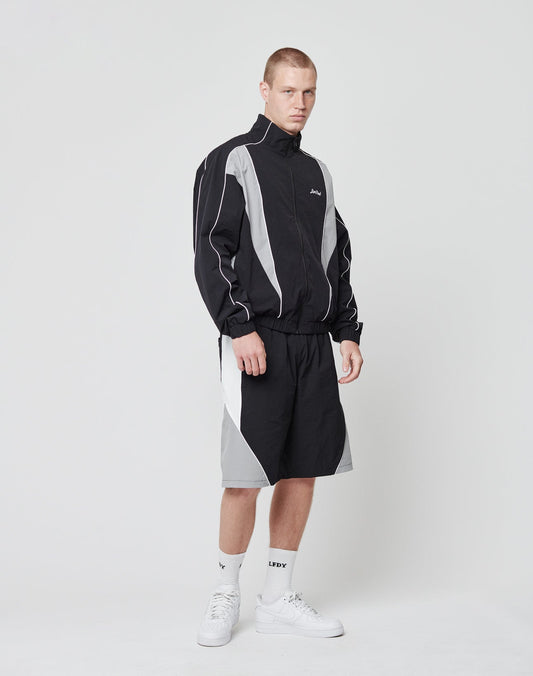 A man stands in a studio wearing the LIVE FAST DIE YOUNG Basic Nylon Trackjacket with matching shorts, white socks, and sneakers. He faces the camera with a neutral expression against a plain white background.