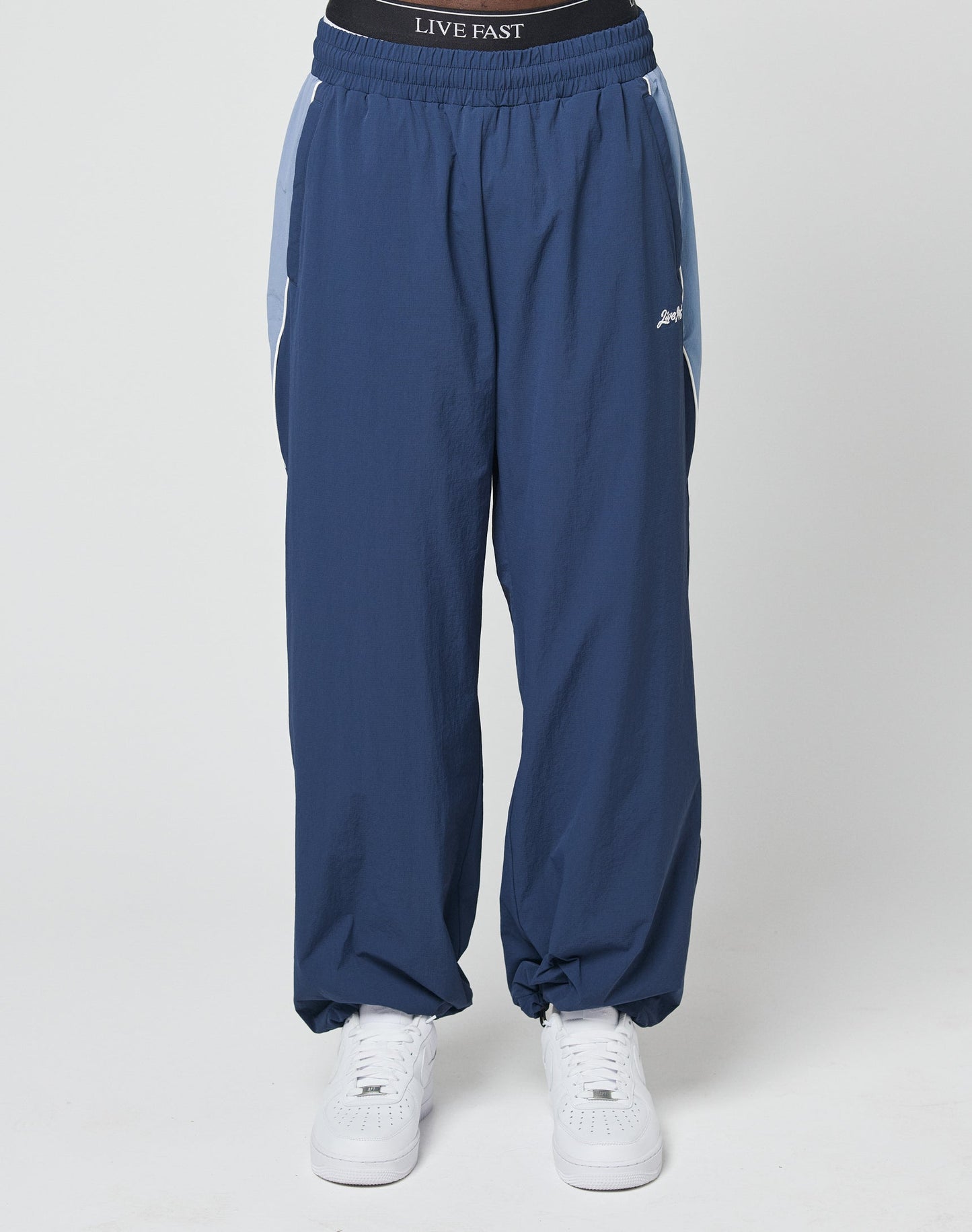 A person wearing LIVE FAST DIE YOUNG Basic Nylon Trackpants in wide fit blue with white side stripes and an Elastikbund waistband that says LIVE FAST, paired with white sneakers, stands against a plain white background.