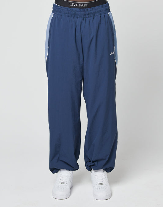 A person wearing LIVE FAST DIE YOUNG Basic Nylon Trackpants in wide fit blue with white side stripes and an Elastikbund waistband that says LIVE FAST, paired with white sneakers, stands against a plain white background.
