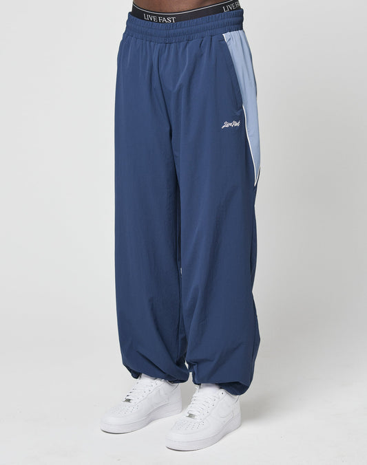 A person wears LIVE FAST DIE YOUNG Basic Nylon Trackpants in wide fit blue with an elastic waistband labelled Live Fast. Light blue side panels and zip pockets add style. The look is finished with white sneakers against a plain white background.