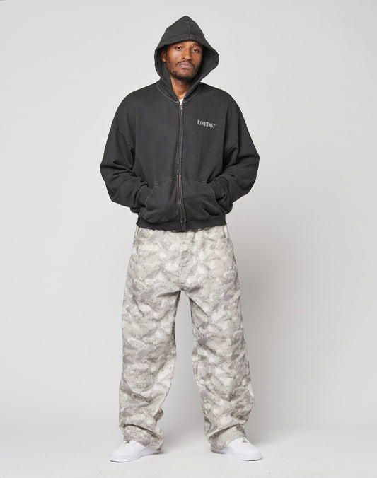 A man wears the LIVE FAST DIE YOUNG LFDY Loyal Hooded Zip, a black vintage-washed, oversize-fit hoodie, paired with gray camo baggy pants and white sneakers, standing against a plain light background with hands in his pockets.