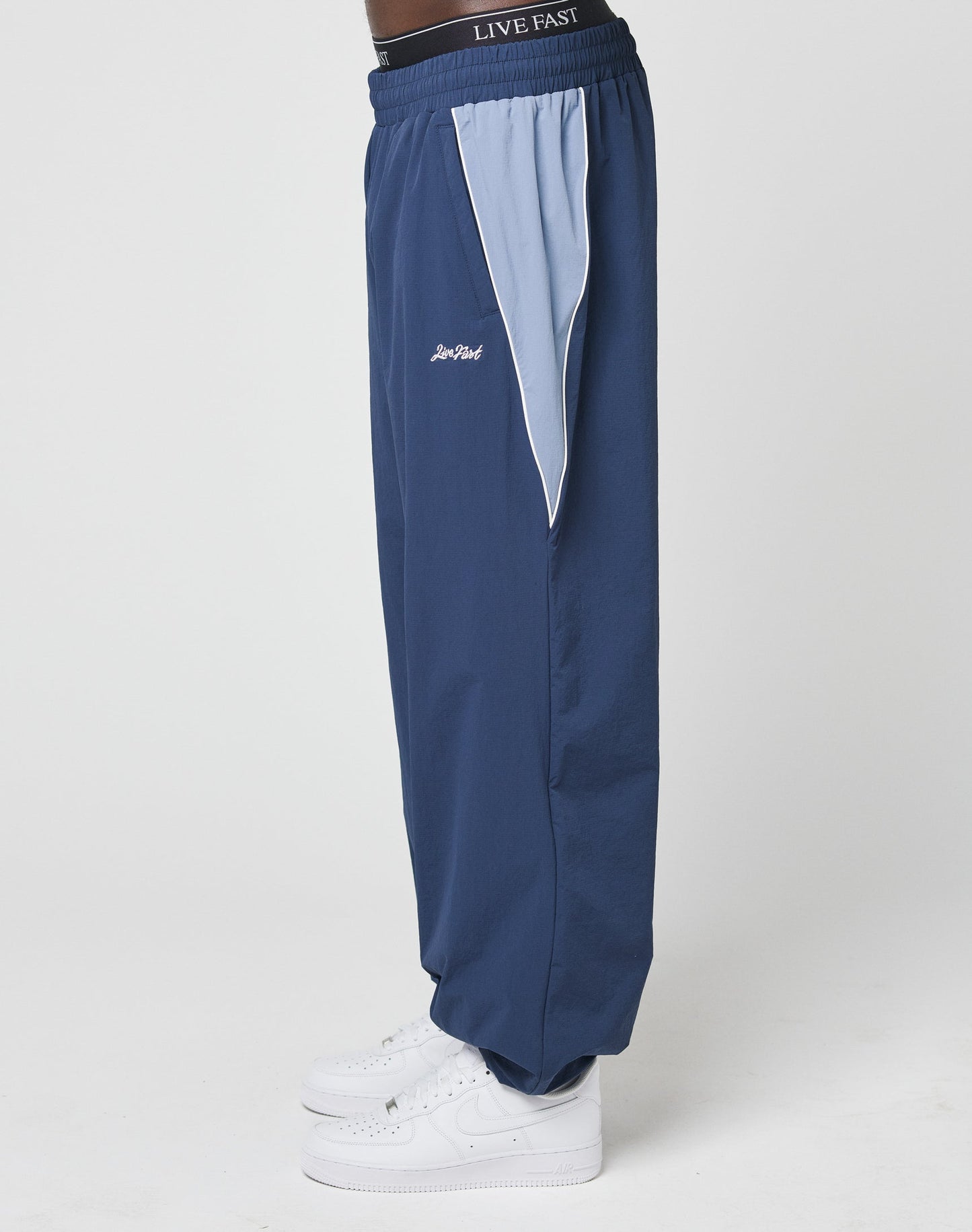 A person wears the LIVE FAST DIE YOUNG Basic Nylon Trackpants in blue with light blue and white side panels, white sneakers, and an elastic waistband featuring LIVE FAST text, standing against a plain white background.