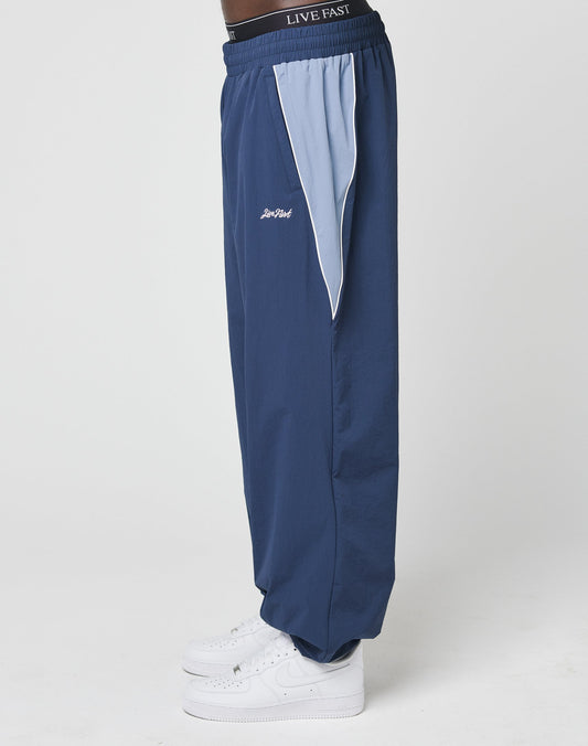 A person wears the LIVE FAST DIE YOUNG Basic Nylon Trackpants in blue with light blue and white side panels, white sneakers, and an elastic waistband featuring LIVE FAST text, standing against a plain white background.