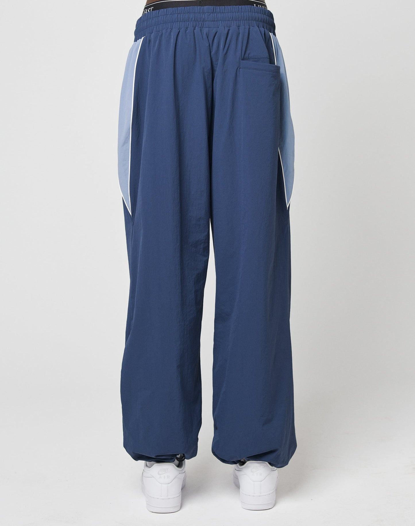 A person wearing LIVE FAST DIE YOUNG Basic Nylon Trackpants, wide fit in navy blue with light blue side panels and an elastic waistband, paired with white sneakers, photographed from the back against a plain white background.