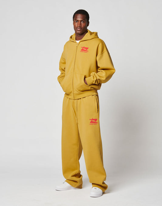 A man stands against a white background wearing the LIVE FAST DIE YOUNG Live Fast Sweatsuit Jogger in mustard yellow with red graphics, an elastik-bund, and white sneakers.