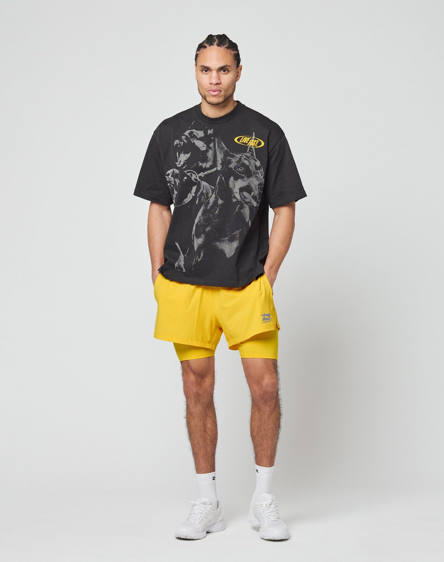 A person with braided hair faces away, wearing the LIVE FAST DIE YOUNG LFDY Bark Tee in black vintage wash and bright yellow oversized shorts, set against a plain white background.