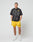 A man stands against a plain background wearing the oversized black LFDY Bark Tee by LIVE FAST DIE YOUNG, teamed with bright yellow shorts and white athletic shoes. His hands are in his pockets and he looks forward.