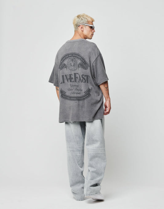 A person with short blond hair, sunglasses, and light-wash baggy jeans models the LIVE FAST DIE YOUNG LFDY Loyal Tee in oversize gray with LIVE FAST text on the back, standing against a plain white background.