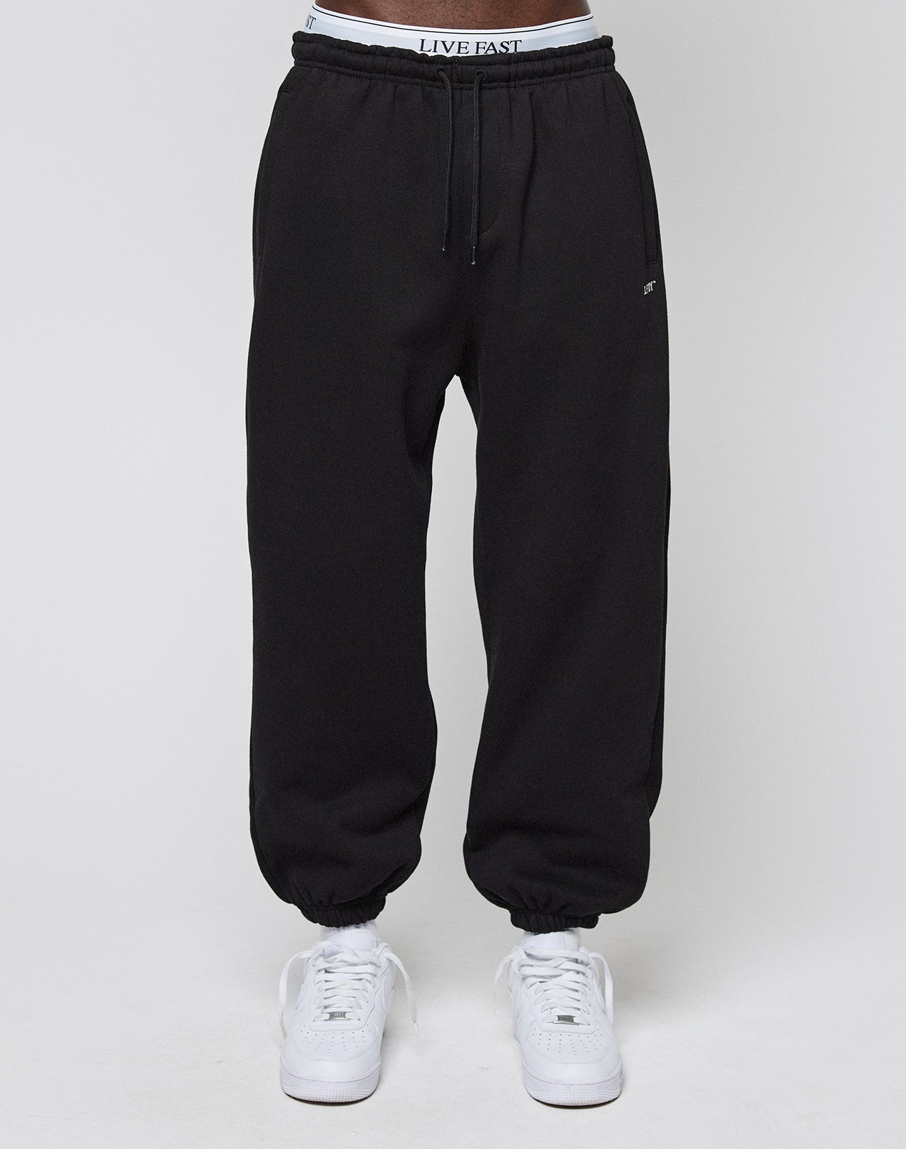 A person wears the Basic Loose Jogger by LIVE FAST DIE YOUNG, featuring an Elastik-Bund, loose fit, and a LIVE FAST logo on the waistband. Paired with white sneakers against a plain white background.