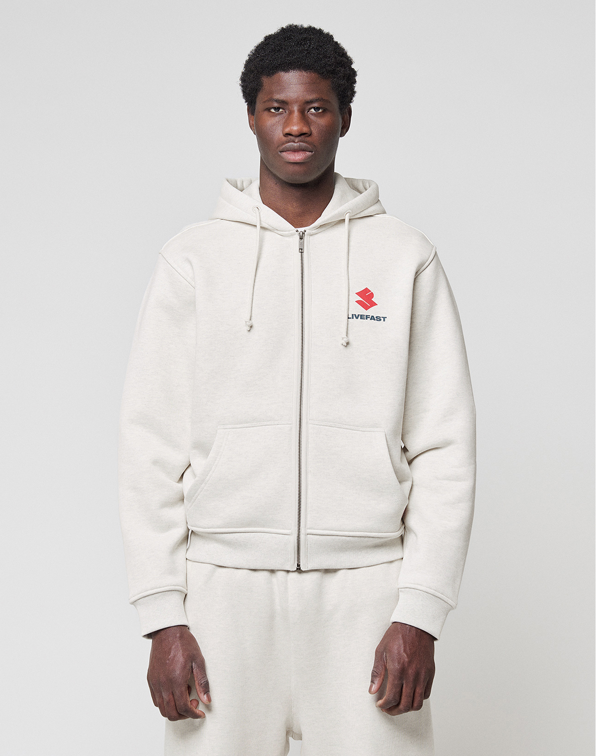 A person in a light beige LFDY Heavyweight Lounge Zip Hooded by LIVE FAST DIE YOUNG and matching sweatpants faces forward. The regular fit hoodie has a small red logo and “LIVEFAST” text on the left chest. Background is plain and light-colored. | model