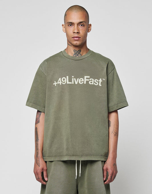 A person with a shaved head and tattoos wears a LIVE FAST DIE YOUNG Basic +49 Tee in olive green, made from 100% Baumwolle, featuring +49 LiveFast on the front, standing against a plain light background.