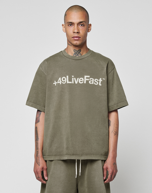 A person with a shaved head and visible tattoos wears the LIVE FAST DIE YOUNG Basic +49 Tee—an oversized olive-green shirt featuring a bold white "+49 Live Fast" front print—while standing against a plain light background. | model