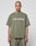 A person with a shaved head and tattoos wears a LIVE FAST DIE YOUNG Basic +49 Tee in olive green, made from 100% Baumwolle, featuring +49 LiveFast on the front, standing against a plain light background.