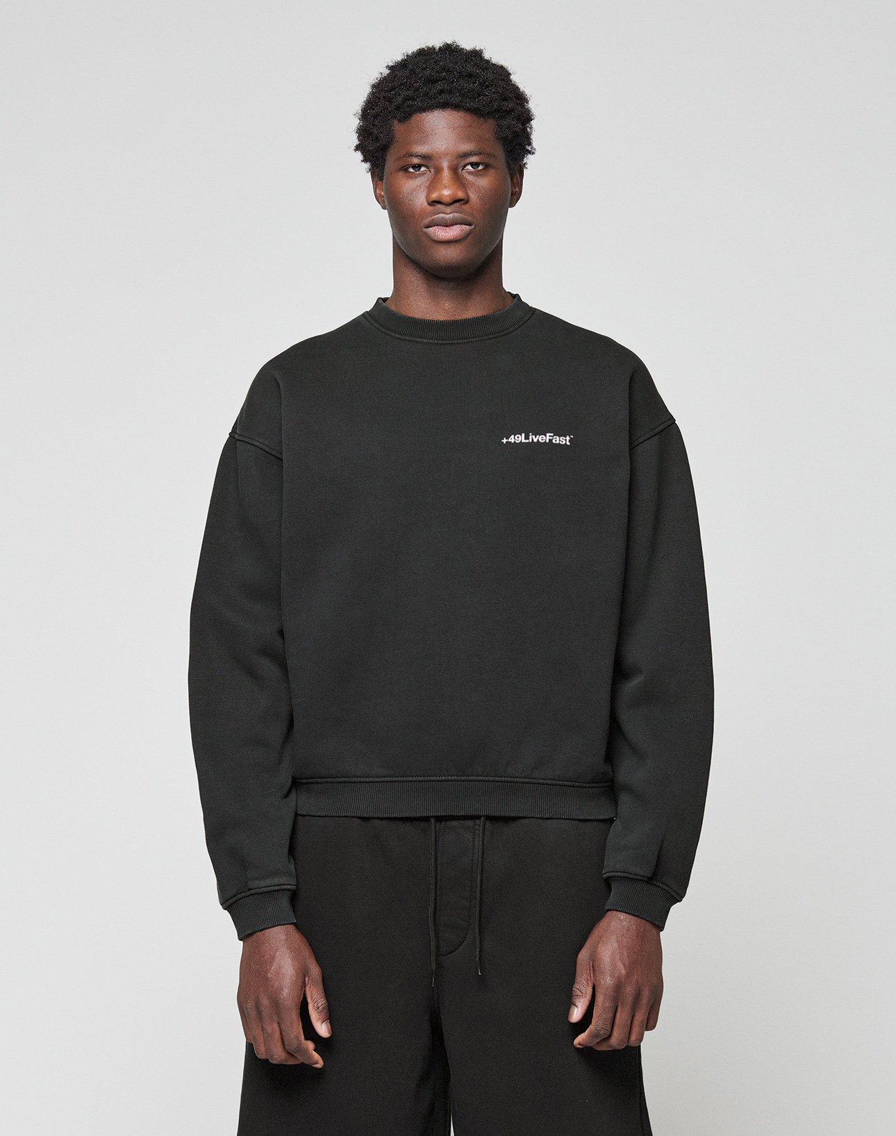 A young man with short curly hair wears the LIVE FAST DIE YOUNG Basic +49 Chest Sweater in black and black pants, standing against a plain light gray background. The sweater features small white #LiveFast text on the left chest. | model