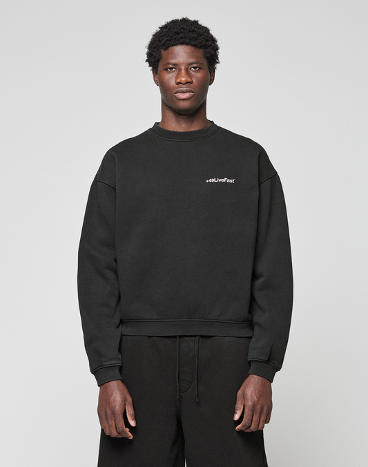 A young man with short curly hair wears the LIVE FAST DIE YOUNG Basic +49 Chest Sweater in black and black pants, standing against a plain light gray background. The sweater features small white #LiveFast text on the left chest. | model