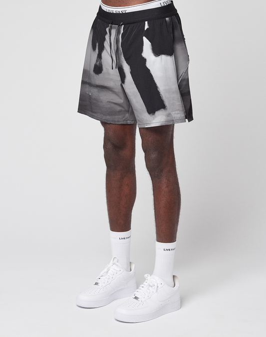 A person stands against a white background wearing LFDY Performance Shorts by LIVE FAST DIE YOUNG with a black-and-white abstract pattern, featuring a reflective logo print. White socks with LIVIN FAST text and white sneakers complete the cropped-below-waist image. | model