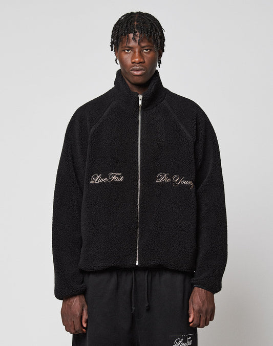 A person wearing a black LFDY Teddy Jacket by LIVE FAST DIE YOUNG, featuring a zipper and “Live Fast” and “Die Young” embroidery on the front, stands against a plain light gray background. | model