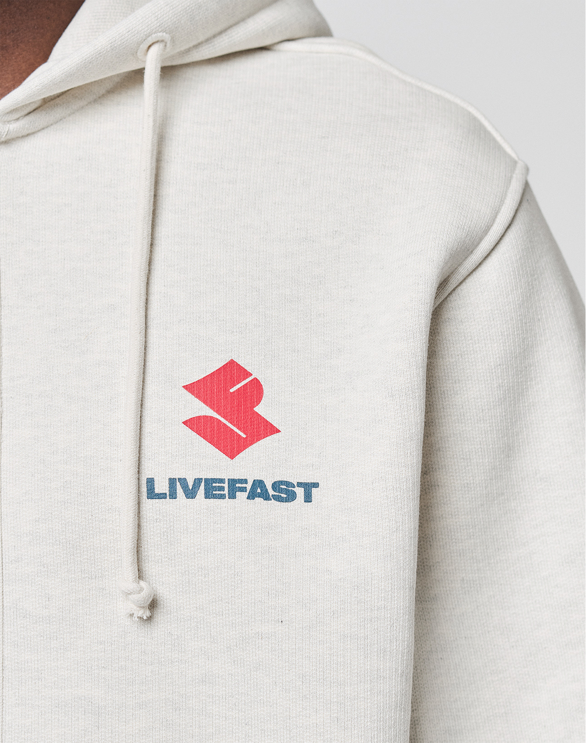 Close-up of the LFDY Heavyweight Lounge Zip Hooded by LIVE FAST DIE YOUNG in light beige, featuring a red abstract logo and blue LIVEFAST text on the left chest. Made from 100% Baumwolle with drawstrings and a comfortable Regular Fit.