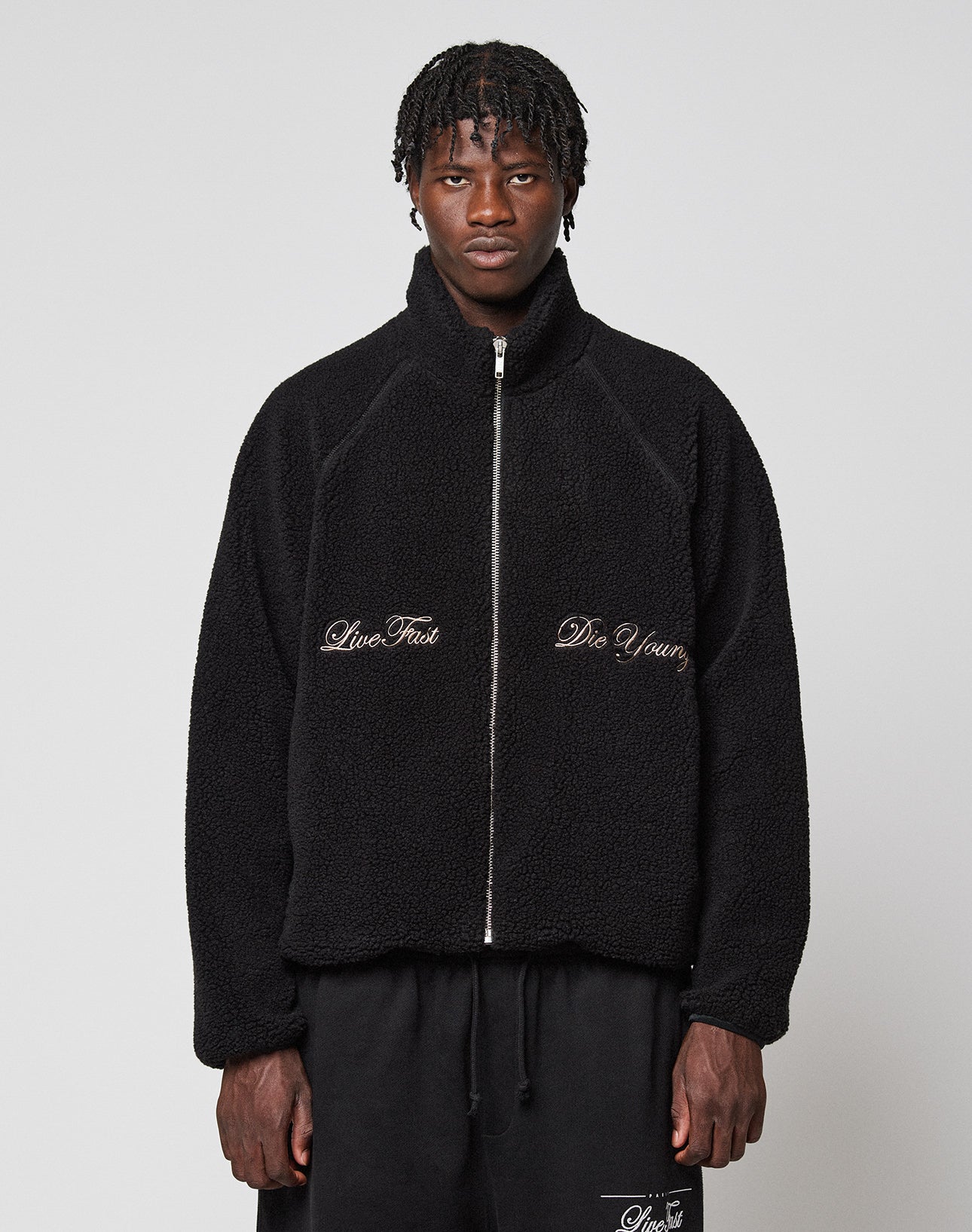 A person wearing a black LFDY Teddy Jacket by LIVE FAST DIE YOUNG, featuring a zipper and “Live Fast” and “Die Young” embroidery on the front, stands against a plain light gray background.