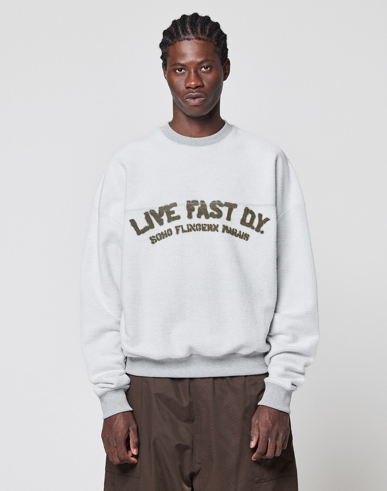 A person with braided hair wears a LIVE FAST DIE YOUNG LFDY District Sweater and loose brown pants, standing against a plain, light background. | model