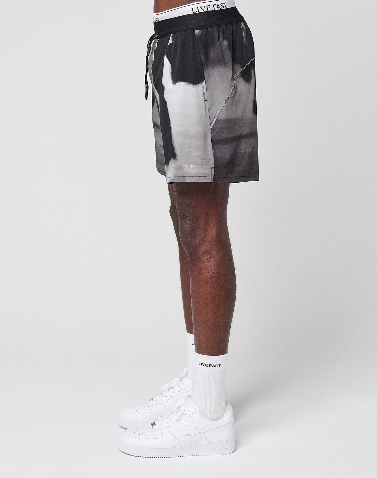 A person stands sideways against a white background, wearing LFDY Performance Shorts by LIVE FAST DIE YOUNG, featuring black and white graphics, a reflective logo print, a white LIVE FAST waistband, socks, and white Nike sneakers.