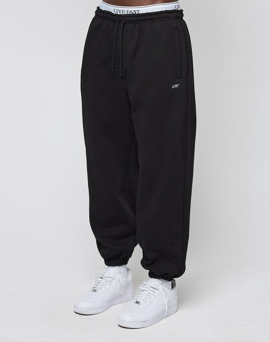 A person wears the LIVE FAST DIE YOUNG Basic Loose Jogger, featuring a loose fit, elastic ankles, and a waistband with LIVE FAST in black lettering, paired with white sneakers against a plain white background.