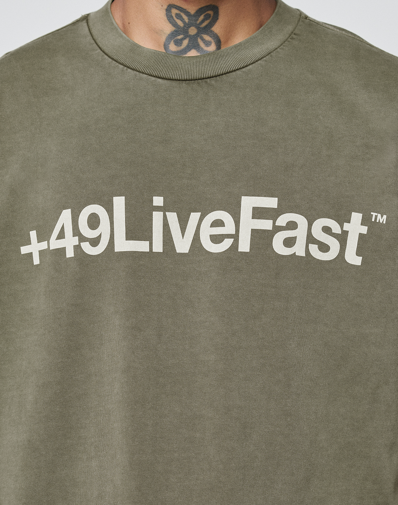 A person wears the LIVE FAST DIE YOUNG Basic +49 Tee, an olive green oversized 100% Baumwolle shirt with a white +49LiveFast™ front print. The collar slightly reveals their neck tattoo.