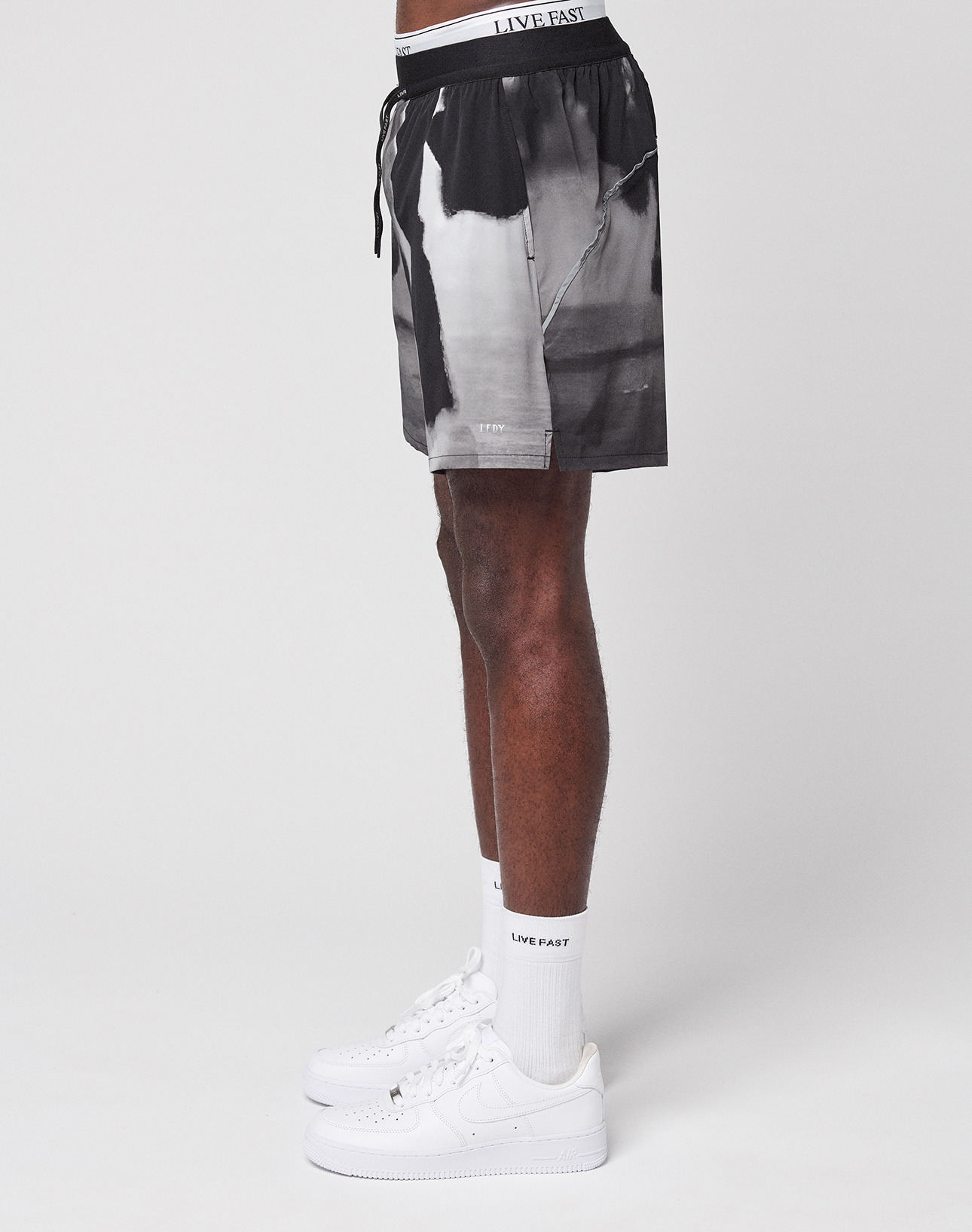 A person stands sideways against a white background, wearing LFDY Performance Shorts by LIVE FAST DIE YOUNG, featuring black and white graphics, a reflective logo print, a white LIVE FAST waistband, socks, and white Nike sneakers.