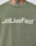 Someone wears an olive green Basic +49 Tee by LIVE FAST DIE YOUNG, featuring +49LiveFast™ in bold white on the chest and topstitching at the neckline. Part of a neck tattoo shows above the collar.