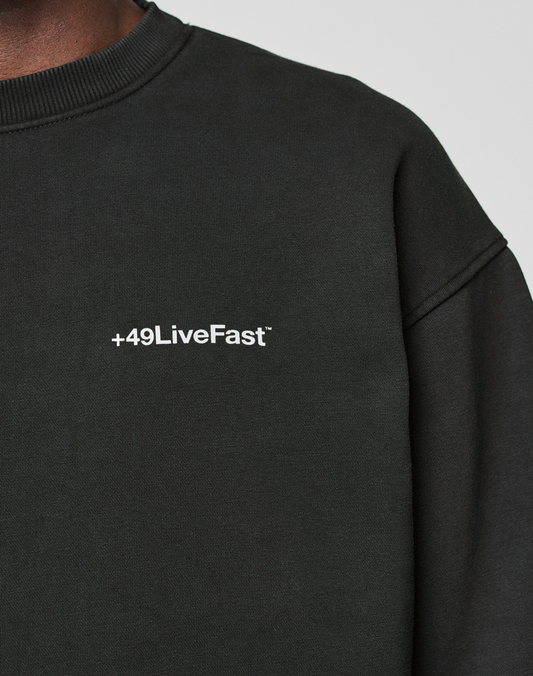 Close-up of a person wearing the LIVE FAST DIE YOUNG Basic +49 Chest Sweater in black, featuring a white "+49" chest print. Only part of the neck, shoulder, and upper chest is visible.