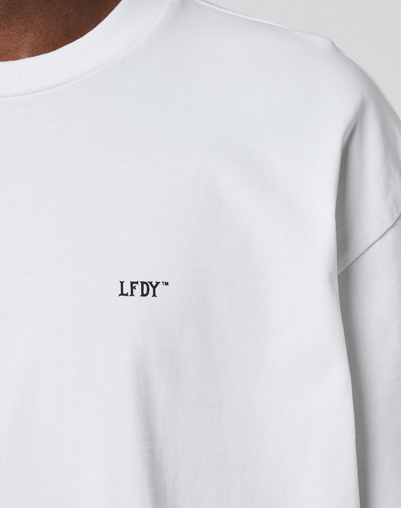 Close-up of a person wearing the LFDY Monaco Boys Tee by LIVE FAST DIE YOUNG in an Oversize Fit, featuring black LFDY™ text on the left chest. Only part of the shirt plus the neck and shoulder are visible.