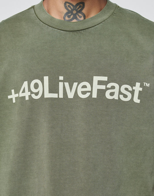 Someone wears an olive green Basic +49 Tee by LIVE FAST DIE YOUNG, featuring +49LiveFast™ in bold white on the chest and topstitching at the neckline. Part of a neck tattoo shows above the collar.
