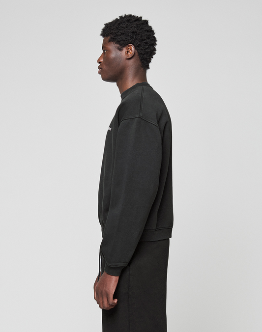 A person with short curly hair stands in profile against a plain light background, wearing the LIVE FAST DIE YOUNG Basic +49 Chest Sweater—an oversize fit black long-sleeve sweatshirt made from Baumwolle Polyester, paired with black pants.