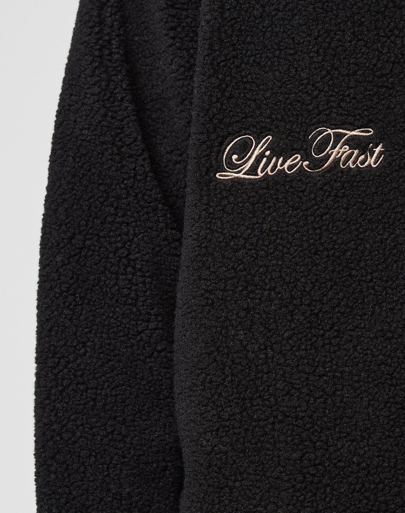 Close-up of the LFDY Teddy Jacket by LIVE FAST DIE YOUNG, featuring black textured fleece, raglan sleeves, and "Live Fast" embroidered in cursive with light-colored thread.
