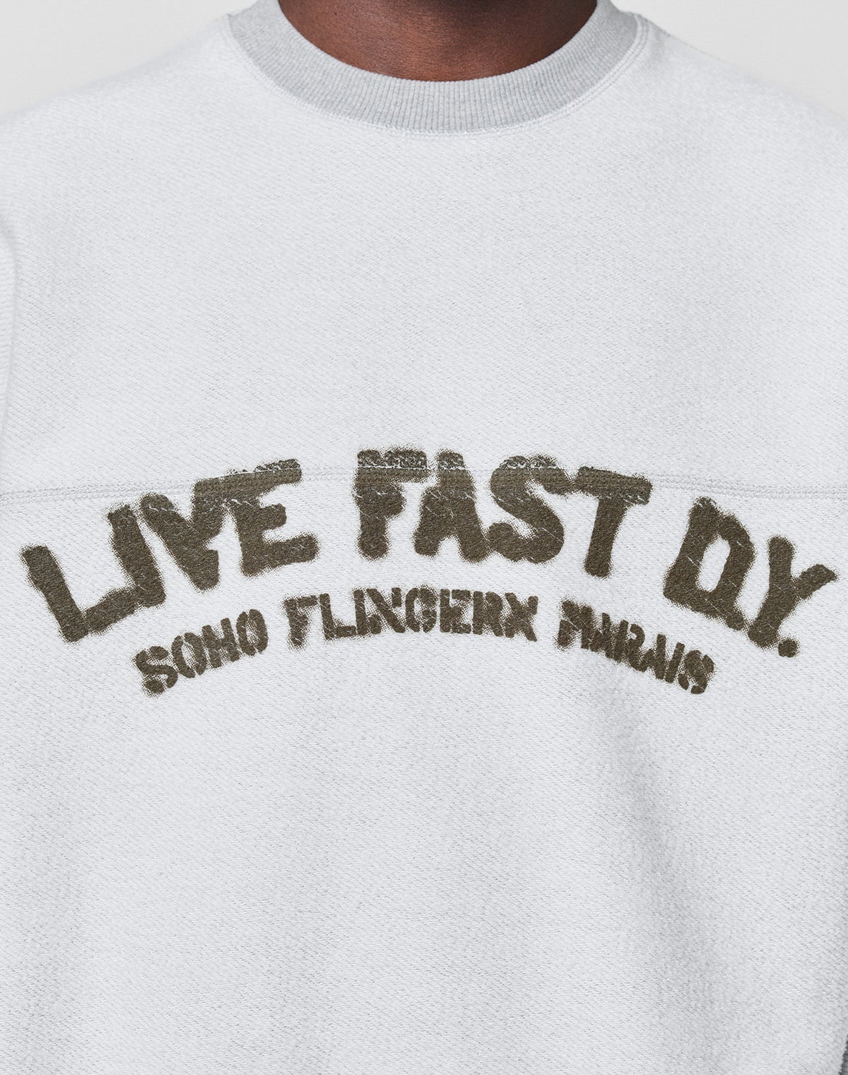 A person wears the LFDY District Sweater by LIVE FAST DIE YOUNG—an oversized white textured sweatshirt in soft cotton-polyester, featuring "LIVE FAST D*Y. SOHO FLINGERN PARIS" in large, distressed letters on the chest.