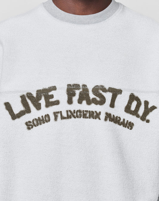 A person wears the LFDY District Sweater by LIVE FAST DIE YOUNG—an oversized white textured sweatshirt in soft cotton-polyester, featuring "LIVE FAST D*Y. SOHO FLINGERN PARIS" in large, distressed letters on the chest.