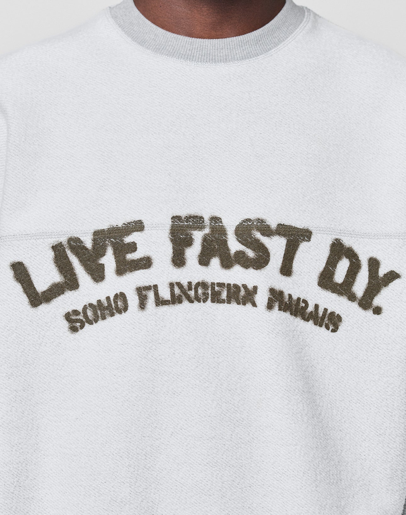 A person wears the LFDY District Sweater by LIVE FAST DIE YOUNG—an oversized white textured sweatshirt in soft cotton-polyester, featuring "LIVE FAST D*Y. SOHO FLINGERN PARIS" in large, distressed letters on the chest.