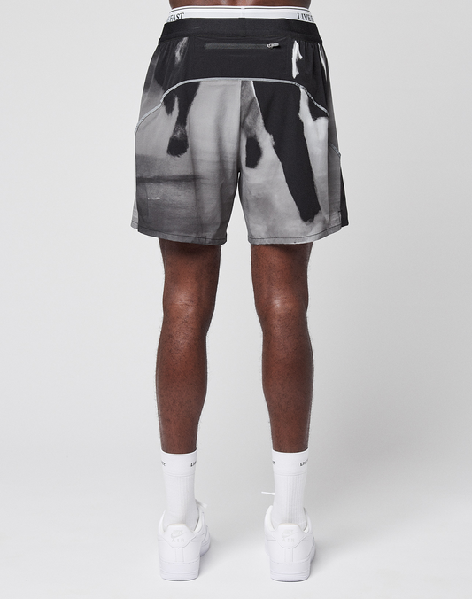 A person stands facing away, showing only their lower half, wearing LIVE FAST DIE YOUNG LFDY Performance Shorts with a blurry black and white print, paired with white socks and white sneakers featuring a reflective logo print, against a plain light background.