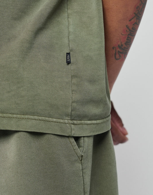 A close-up shows a person in the LIVE FAST DIE YOUNG Basic +49 Tee, Oversize Fit in olive green. Visible are the side seam, small black tag, part of a tattooed arm, and subtle topstitching at the neckline.