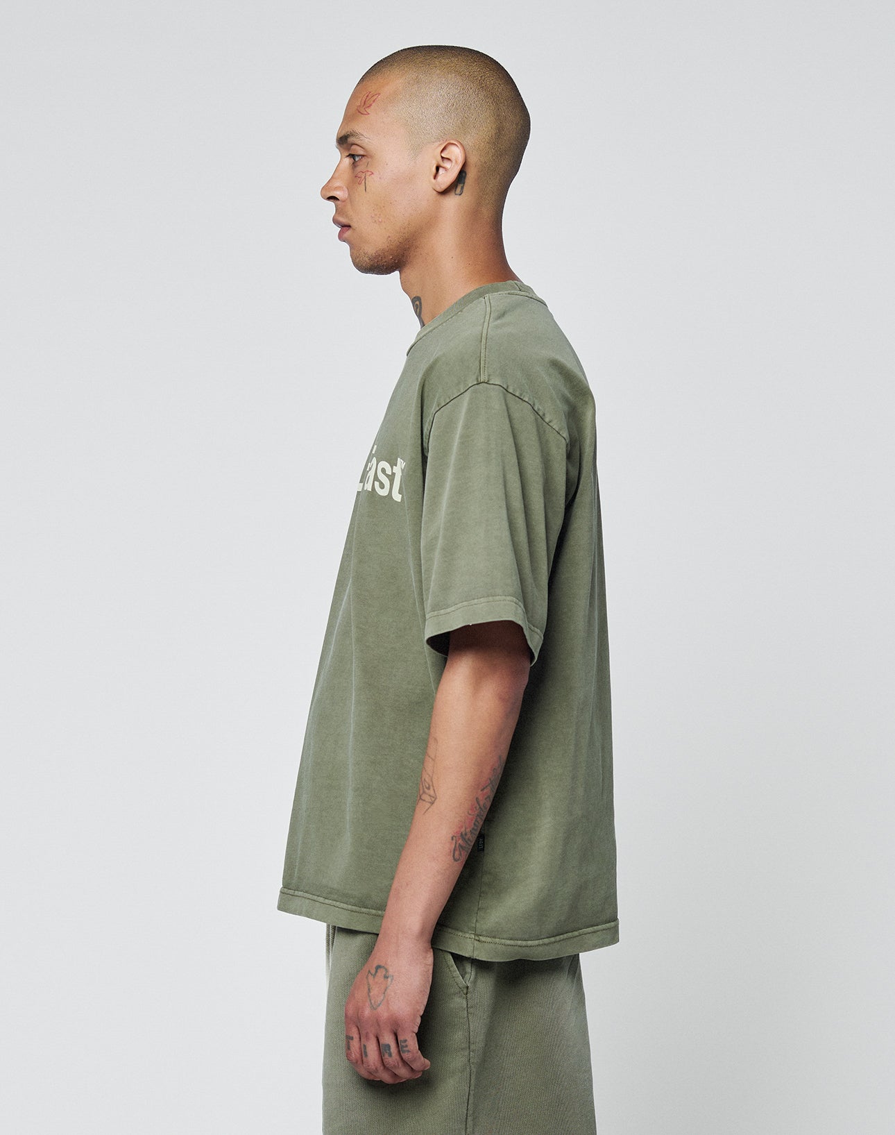 A man with a shaved head, in profile, wears the LIVE FAST DIE YOUNG Basic +49 Tee—an oversized olive-green T-shirt with topstitching at the neckline and matching 100% cotton shorts—against a plain light gray background.