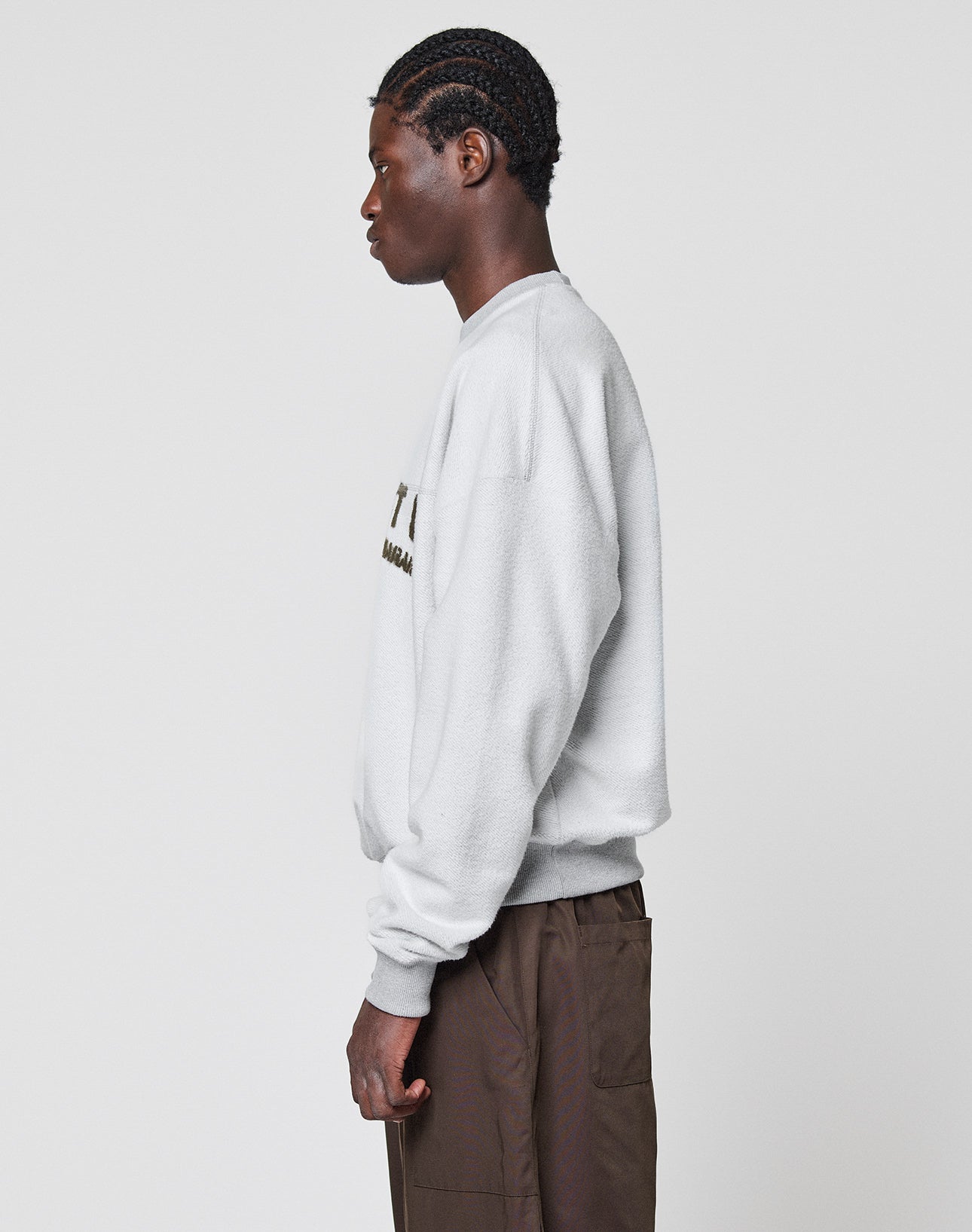 A person with braided hair stands in profile, facing left, wearing the LIVE FAST DIE YOUNG LFDY District Sweater in oversize fit light gray (Baumwolle Polyester, 480 GSM), paired with brown pants, against a plain light background.