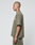 A person with a shaved head stands in profile, facing left, wearing the LIVE FAST DIE YOUNG Basic +49 Tee in olive green with matching shorts. The T-shirt features an oversize fit and is made from 100% Baumwolle. The background is plain and light-colored.