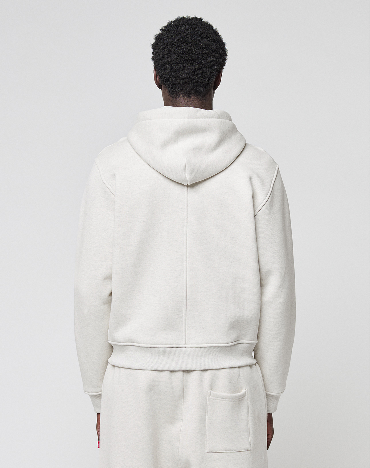A person stands with their back to the camera, dressed in the LFDY Heavyweight Lounge Zip Hooded by LIVE FAST DIE YOUNG and matching sweatpants, both off-white and made from 100% Baumwolle, against a plain light background.