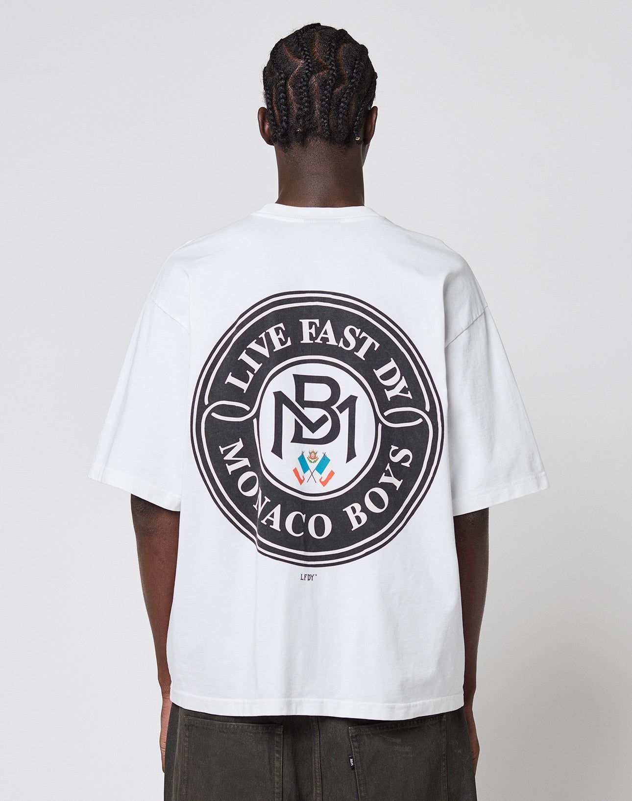 Seen from the back, a person with braided hair wears the LFDY Monaco Boys Tee by LIVE FAST DIE YOUNG—an oversized white 100% Baumwolle shirt with a large circular black graphic reading LIVE FAST BY MONACO BOYS and MB1 in the center.