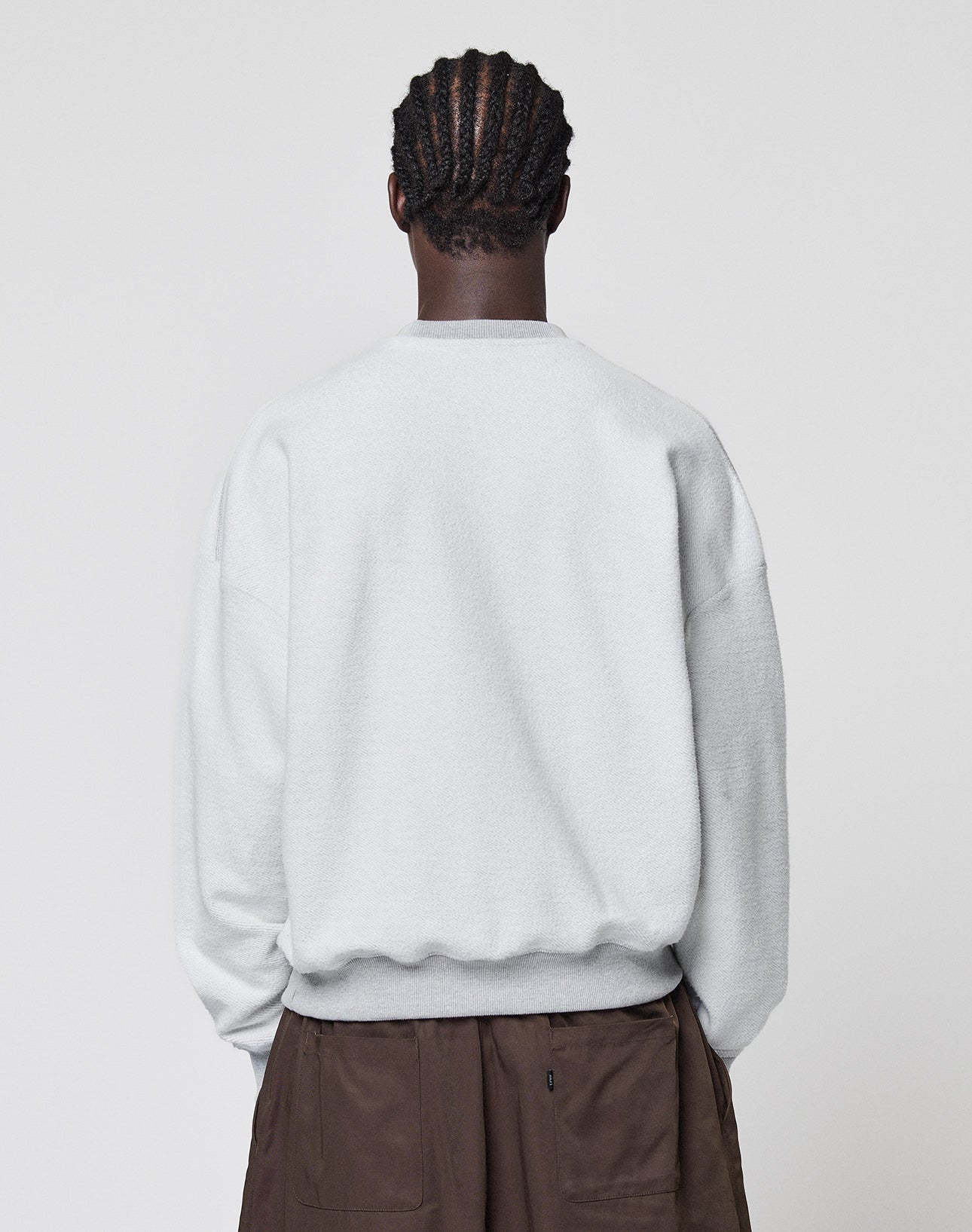 A person with cornrow braids is shown from behind, wearing a light gray LIVE FAST DIE YOUNG LFDY District Sweater and brown pants, standing against a plain light background.