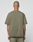 Seen from behind, a person with a shaved head wears the oversized Basic +49 Tee in olive green by LIVE FAST DIE YOUNG, paired with matching shorts. Tattooed arms add edge against a plain light backdrop.