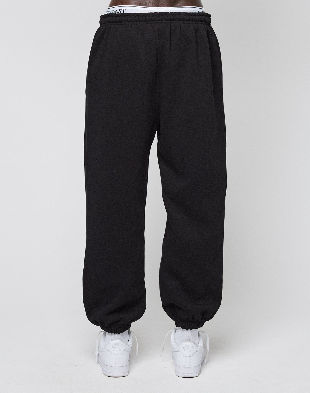 A person wearing LIVE FAST DIE YOUNG Basic Loose Jogger sweatpants in black with an Elastik-Bund and white sneakers, standing against a plain white background.